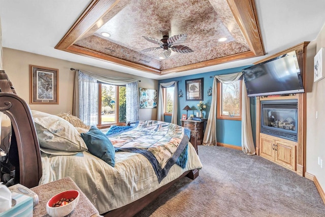 carpeted bedroom featuring baseboards, a raised ceiling, a ceiling fan, and a glass covered fireplace