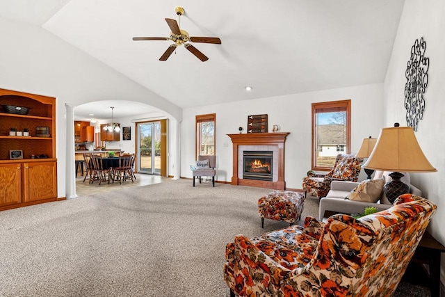 carpeted living area with baseboards, ceiling fan, vaulted ceiling, arched walkways, and a glass covered fireplace