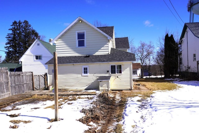 snow covered house featuring a shingled roof and fence