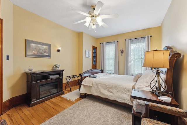 bedroom featuring light wood-style floors, ceiling fan, baseboards, and a glass covered fireplace