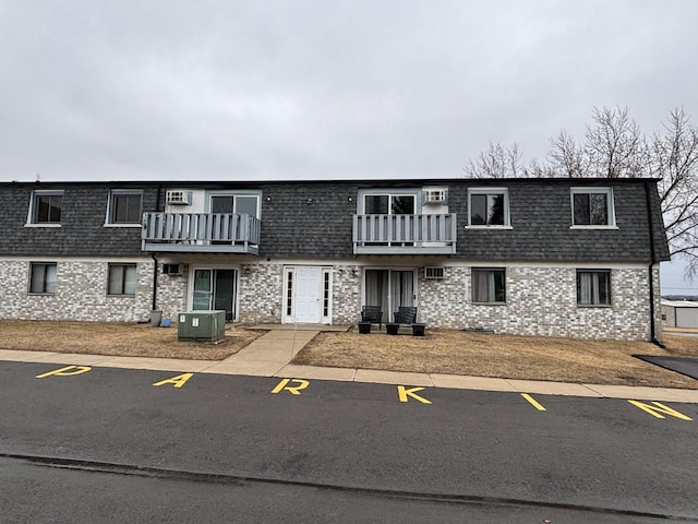 view of front of house featuring mansard roof, a balcony, roof with shingles, central AC, and brick siding