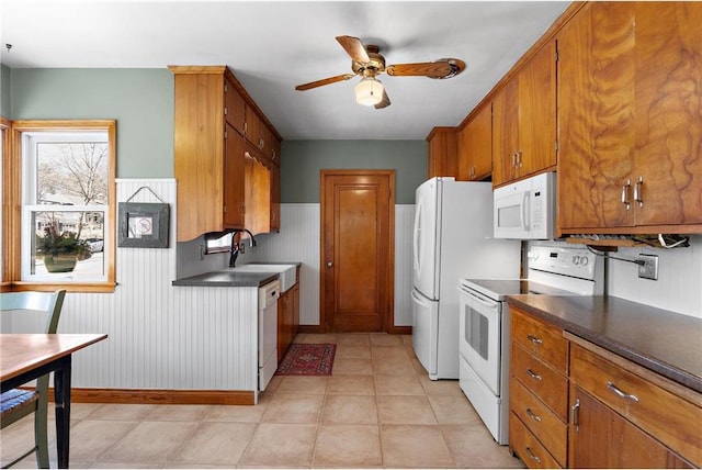 kitchen with white appliances, brown cabinetry, wainscoting, dark countertops, and ceiling fan