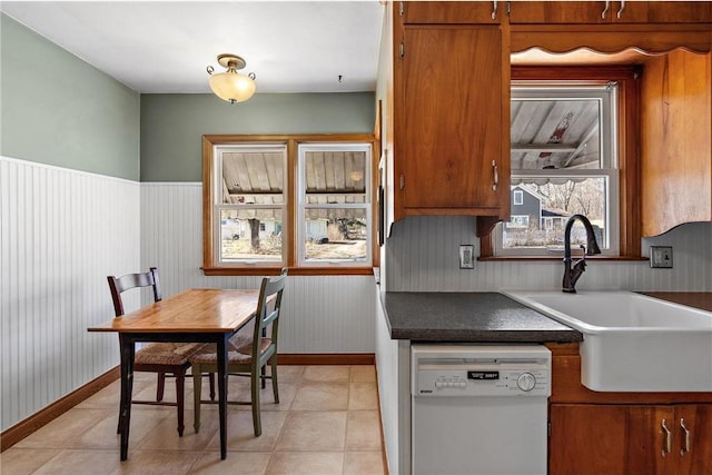 kitchen with brown cabinetry, wainscoting, dark countertops, white dishwasher, and a sink