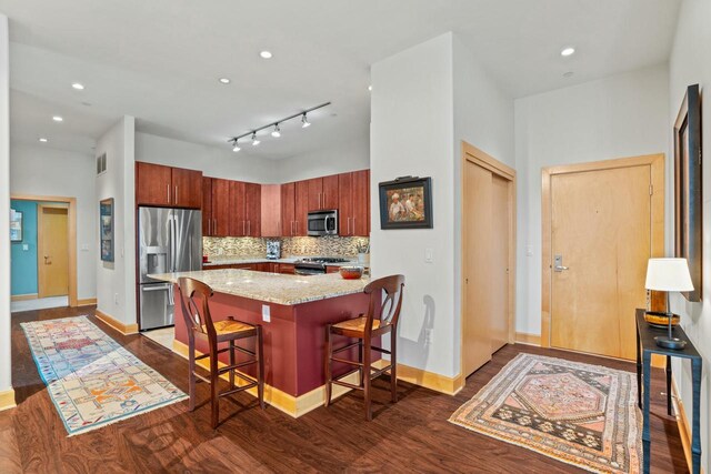 kitchen featuring dark wood-style flooring, backsplash, appliances with stainless steel finishes, light stone countertops, and a kitchen bar