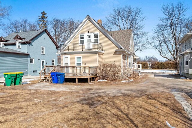 rear view of property featuring dirt driveway, roof with shingles, a chimney, and a balcony