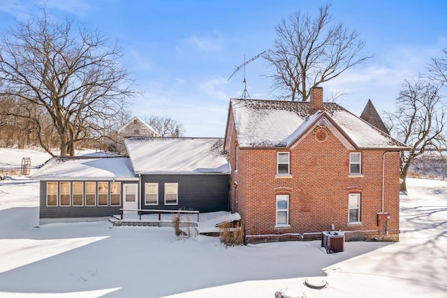 snow covered property with a sunroom, a chimney, central AC unit, and brick siding