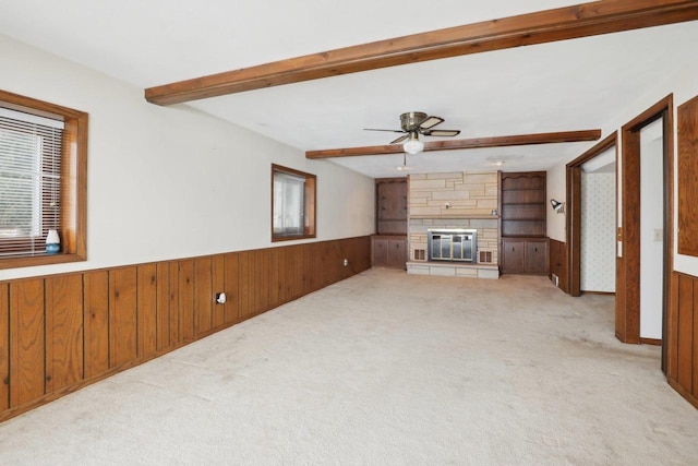 unfurnished living room featuring carpet floors, a wainscoted wall, beamed ceiling, and a stone fireplace