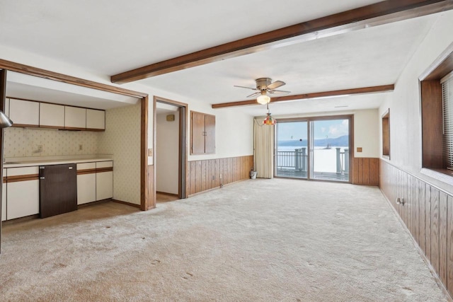 unfurnished living room with a ceiling fan, beam ceiling, wainscoting, and light carpet