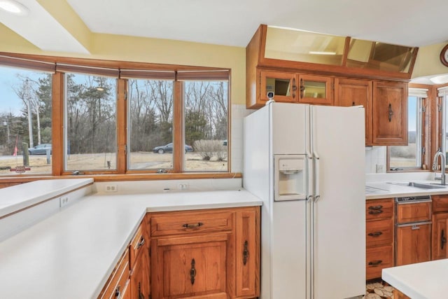 kitchen featuring white refrigerator with ice dispenser, brown cabinetry, glass insert cabinets, light countertops, and a sink