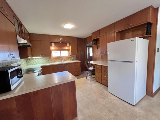 kitchen featuring under cabinet range hood, white appliances, light countertops, decorative backsplash, and brown cabinetry