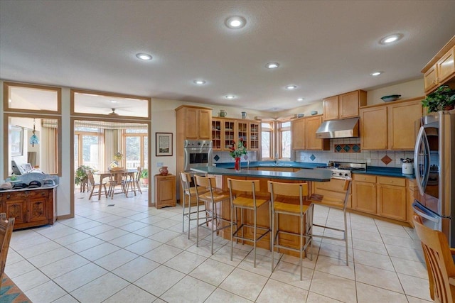 kitchen with light tile patterned floors, stainless steel appliances, under cabinet range hood, dark countertops, and backsplash