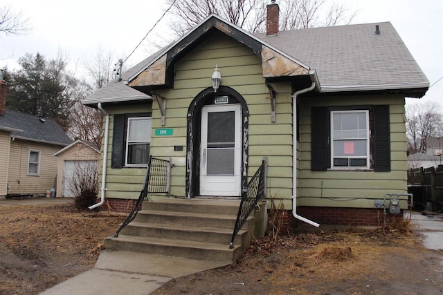 bungalow-style home featuring a garage, an outbuilding, a shingled roof, and a chimney