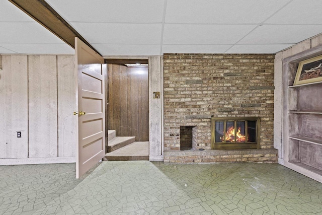 unfurnished living room with built in shelves, a brick fireplace, wood walls, a drop ceiling, and stairs