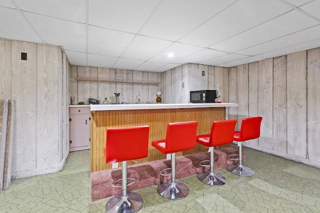 bar with a bar, wooden walls, black microwave, and a paneled ceiling