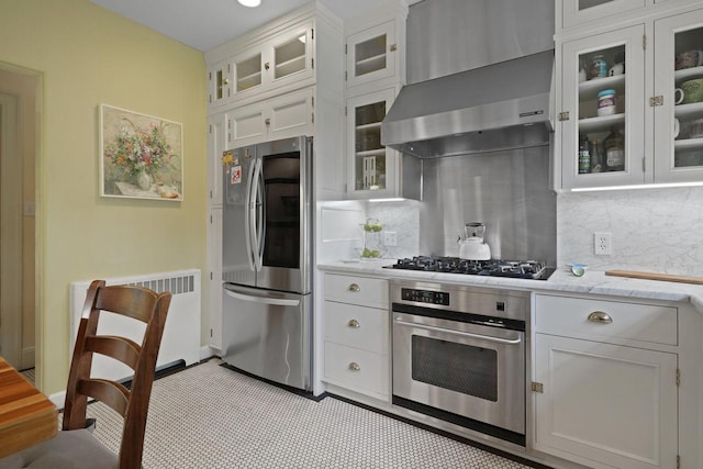 kitchen featuring white cabinetry, appliances with stainless steel finishes, decorative backsplash, wall chimney exhaust hood, and radiator