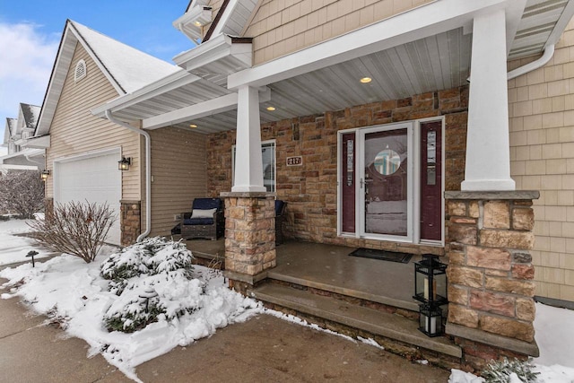 snow covered property entrance featuring a garage, covered porch, and stone siding