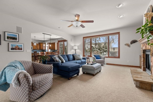 living room with arched walkways, a ceiling fan, light carpet, a stone fireplace, and baseboards