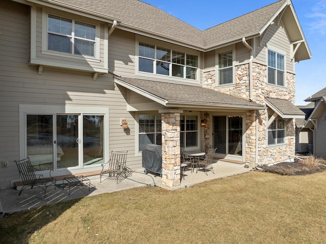 back of house featuring stone siding, roof with shingles, a patio, and a lawn