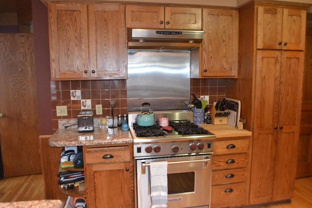 kitchen with light wood finished floors, decorative backsplash, brown cabinetry, high end stainless steel range oven, and under cabinet range hood