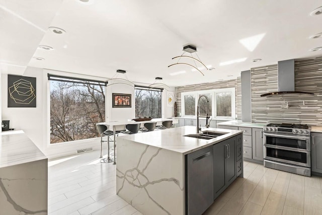 kitchen with stainless steel appliances, gray cabinetry, a healthy amount of sunlight, wall chimney range hood, and a sink