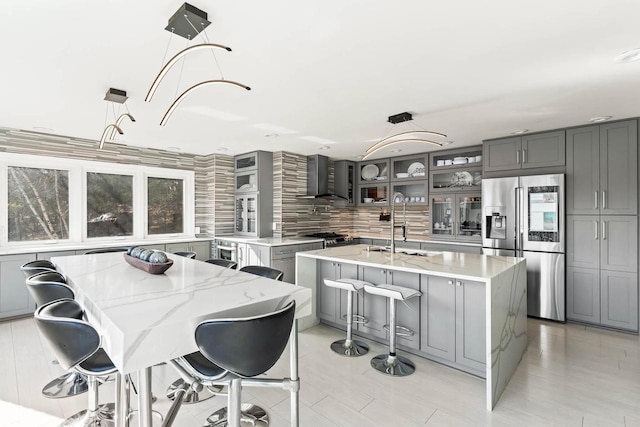 kitchen featuring stainless steel appliances, wall chimney range hood, a center island with sink, and gray cabinetry