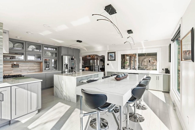 kitchen with a center island with sink, arched walkways, stainless steel fridge with ice dispenser, gray cabinets, and backsplash