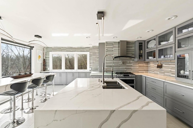 kitchen featuring a sink, gray cabinets, wall chimney exhaust hood, an island with sink, and stainless steel range with gas stovetop