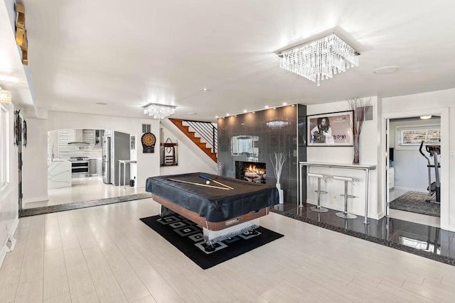 rec room featuring wood finished floors, billiards, baseboards, a lit fireplace, and an inviting chandelier