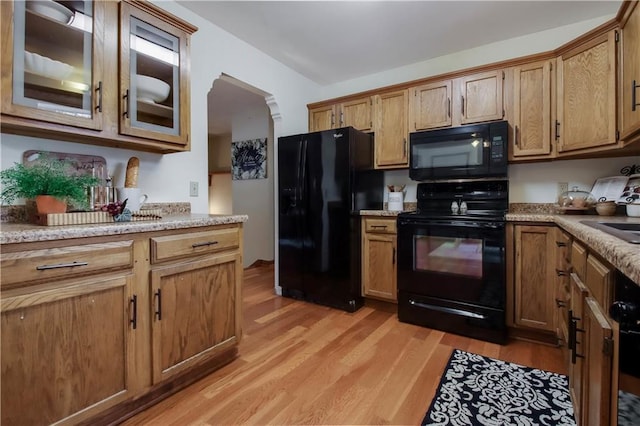 kitchen with brown cabinetry, glass insert cabinets, light wood-style floors, arched walkways, and black appliances