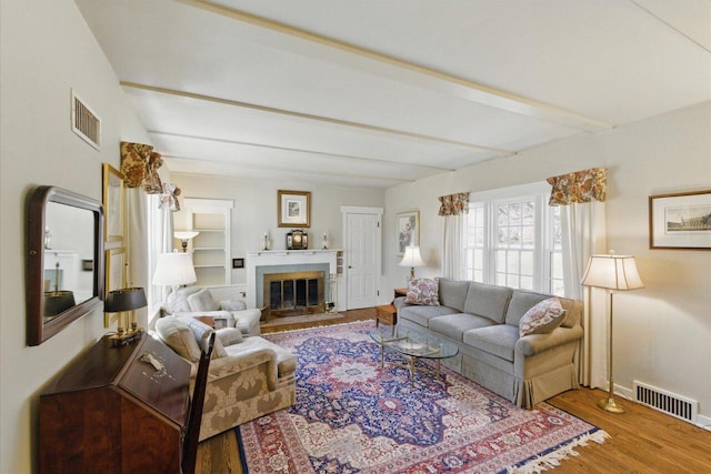 living area featuring a fireplace with flush hearth, beam ceiling, visible vents, and wood finished floors