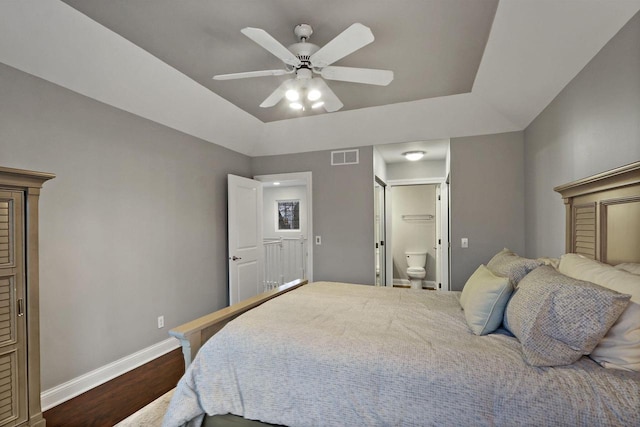 bedroom featuring dark wood-type flooring, visible vents, baseboards, a tray ceiling, and ensuite bath