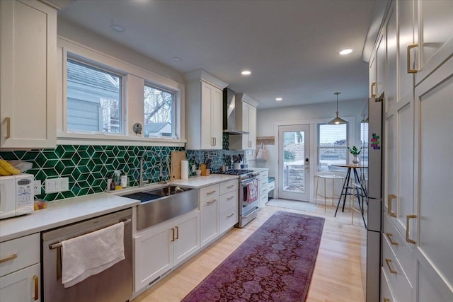 kitchen with light wood-style floors, stainless steel appliances, light countertops, wall chimney range hood, and a sink