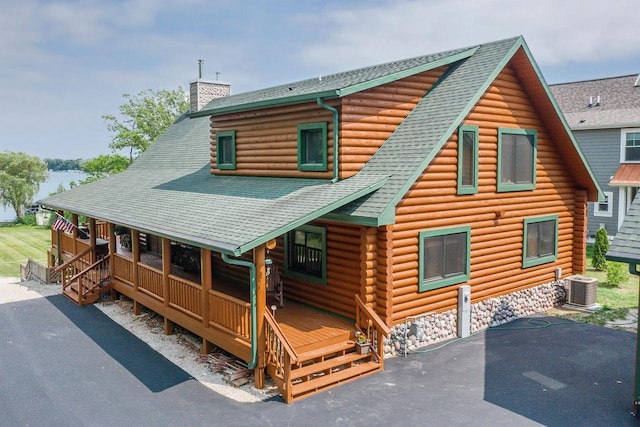 cabin featuring cooling unit, covered porch, a shingled roof, log siding, and a chimney