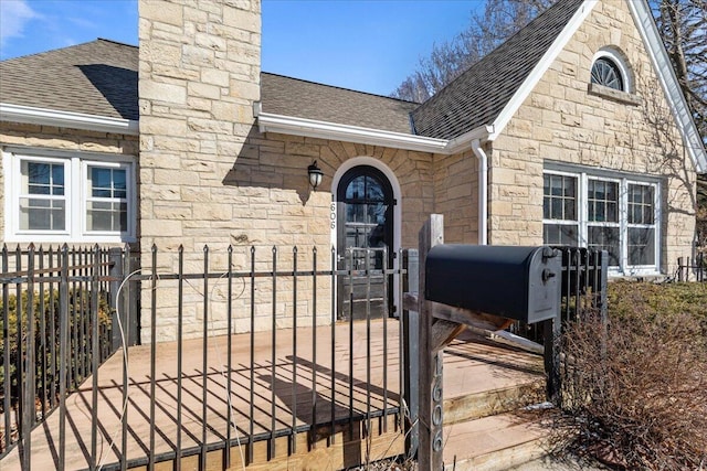 doorway to property with stone siding, a shingled roof, a chimney, and fence