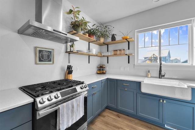 kitchen with blue cabinets, stainless steel gas range, light countertops, wall chimney range hood, and a sink