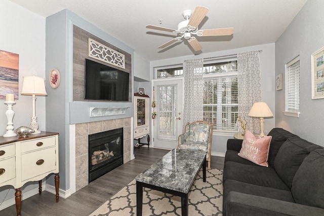 living room with dark wood-style flooring, baseboards, ceiling fan, and a tiled fireplace