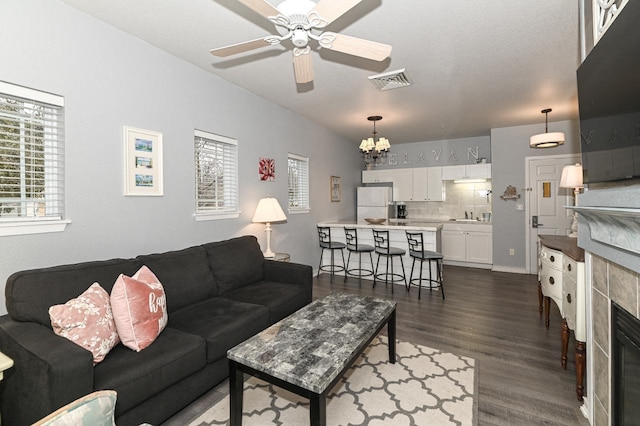 living area with visible vents, dark wood-type flooring, a tiled fireplace, and ceiling fan with notable chandelier