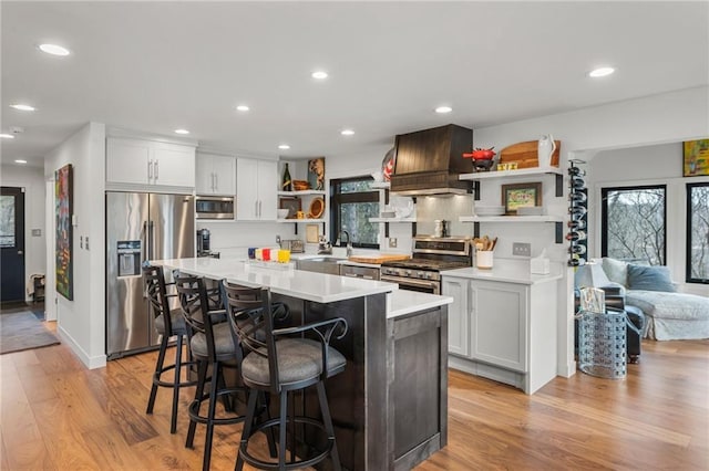 kitchen with light wood-style flooring, light countertops, appliances with stainless steel finishes, open shelves, and custom range hood