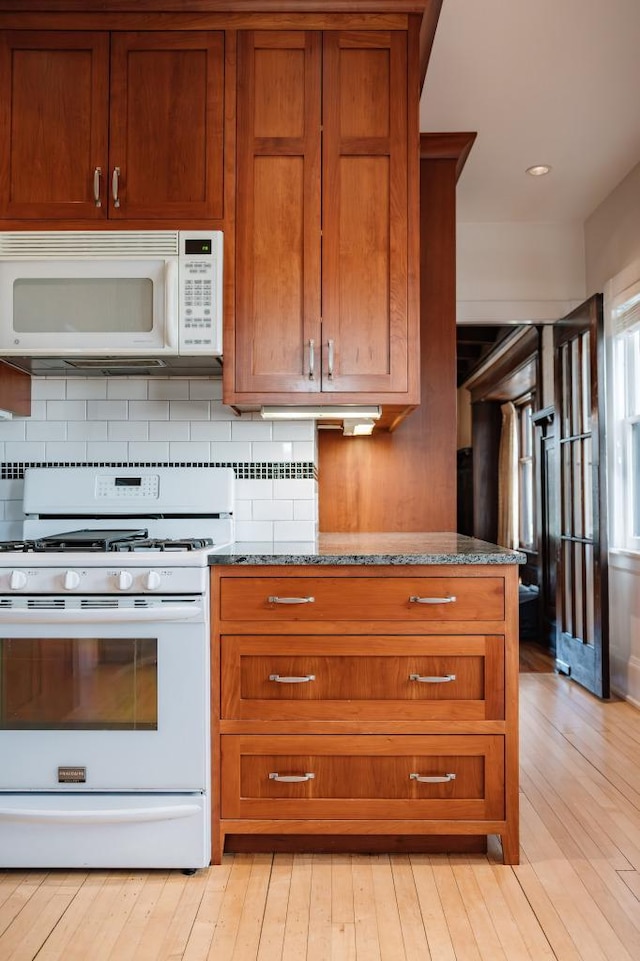 kitchen with light wood-style flooring, white appliances, tasteful backsplash, brown cabinetry, and dark stone countertops