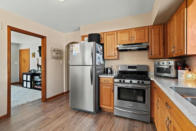 kitchen with arched walkways, under cabinet range hood, stainless steel appliances, wood finished floors, and brown cabinetry