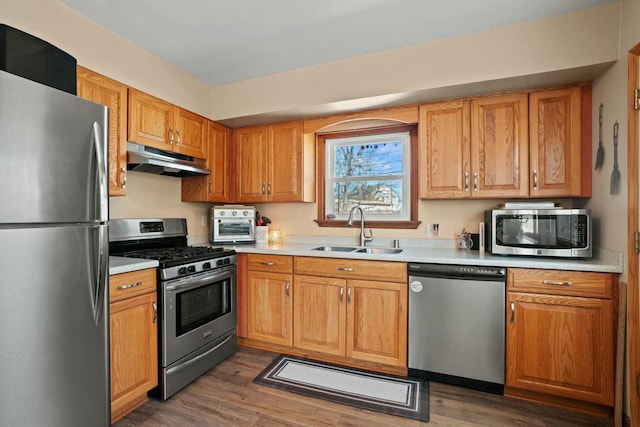 kitchen with appliances with stainless steel finishes, wood finished floors, light countertops, under cabinet range hood, and a sink