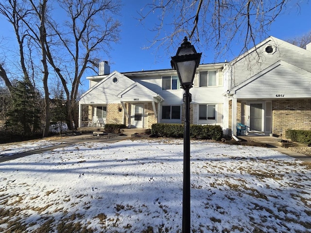 snow covered back of property featuring covered porch, a chimney, and brick siding