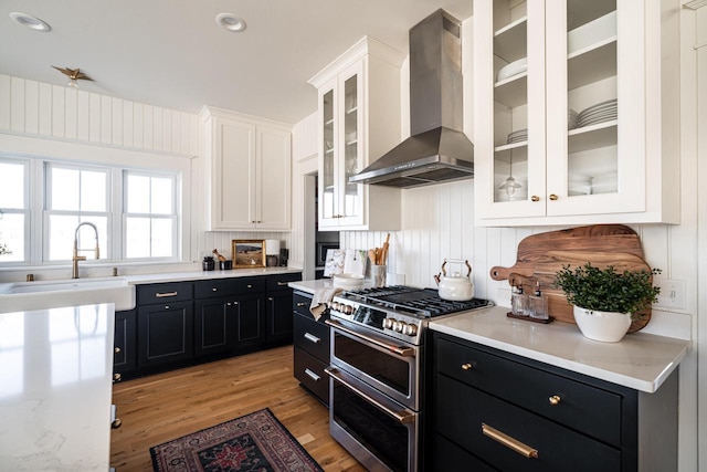 kitchen with double oven range, dark cabinetry, white cabinets, wall chimney exhaust hood, and a sink