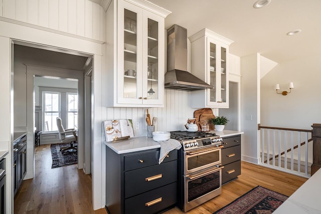 kitchen with light countertops, range with two ovens, light wood-style floors, white cabinetry, and wall chimney exhaust hood