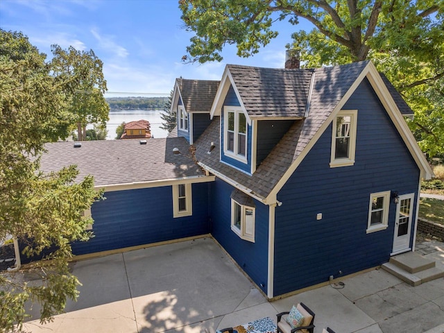 exterior space featuring a patio area, a shingled roof, a chimney, and a water view