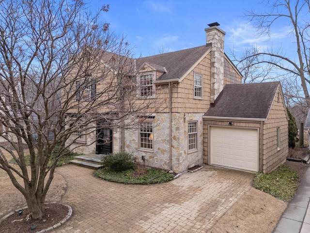 view of front facade with decorative driveway, stone siding, roof with shingles, a garage, and a chimney