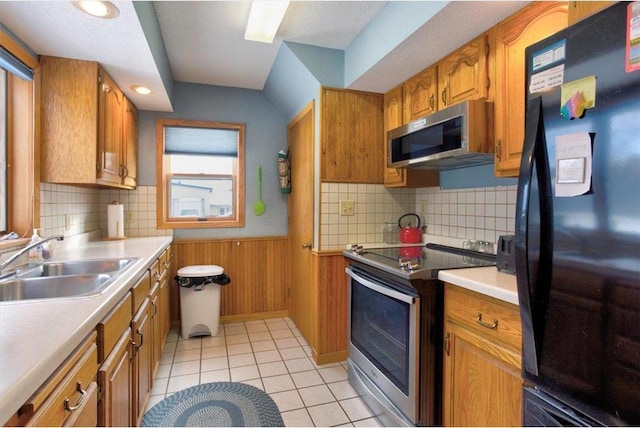 kitchen featuring a sink, stainless steel appliances, wooden walls, brown cabinetry, and wainscoting