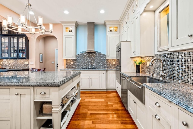 kitchen featuring a sink, open shelves, wall chimney range hood, arched walkways, and light wood finished floors