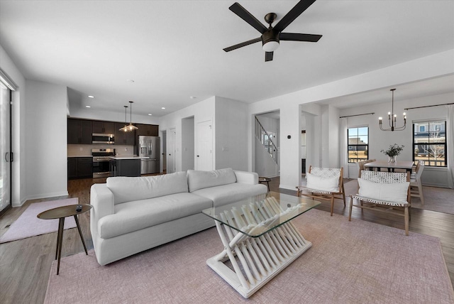 living area featuring stairs, light wood-style flooring, baseboards, and ceiling fan with notable chandelier