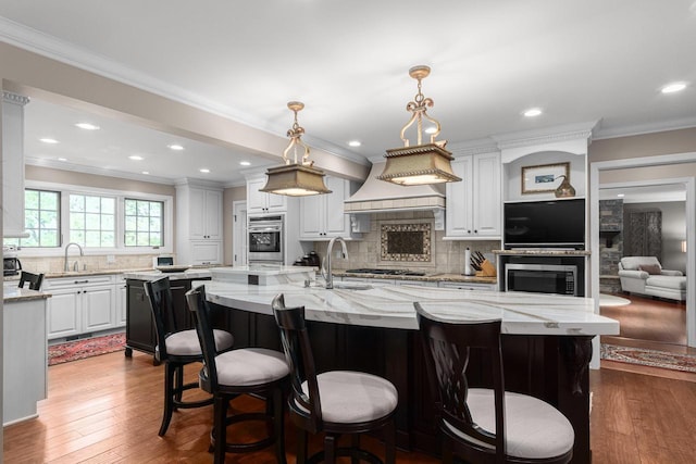 kitchen featuring oven, a center island with sink, custom exhaust hood, white cabinets, and a sink